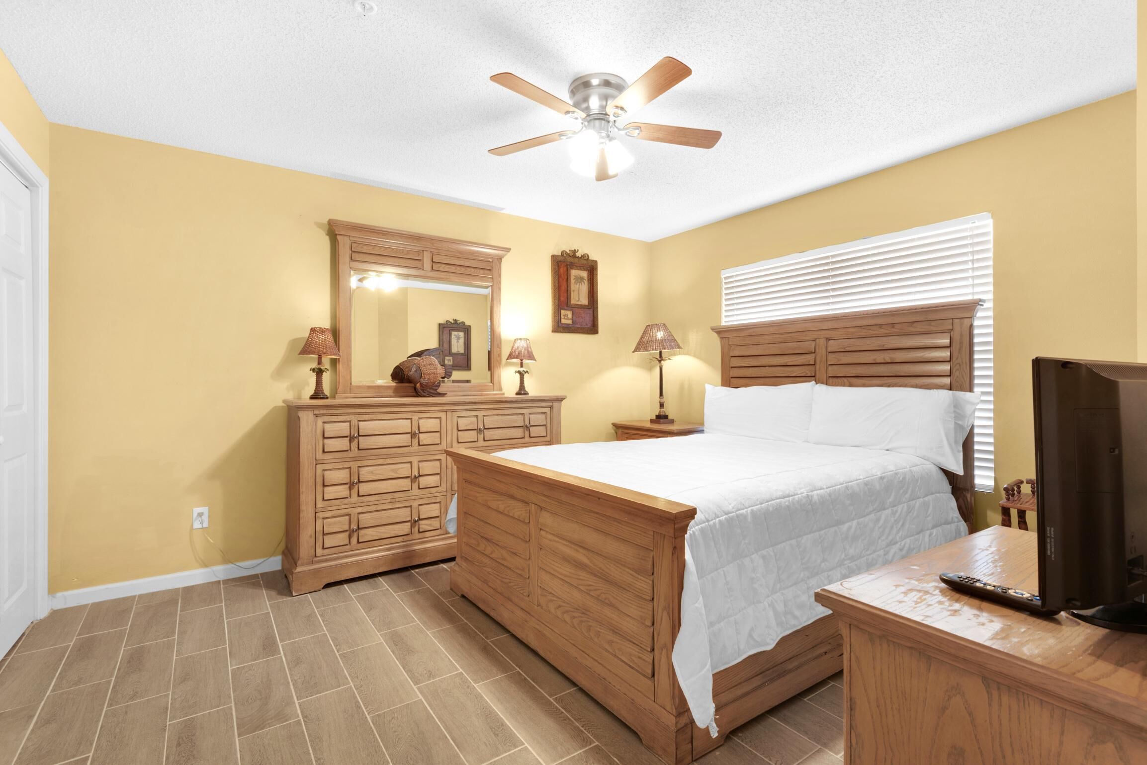 Une chambre à coucher avec un lit en bois, une commode, une coiffeuse avec un miroir et un ventilateur de plafond.