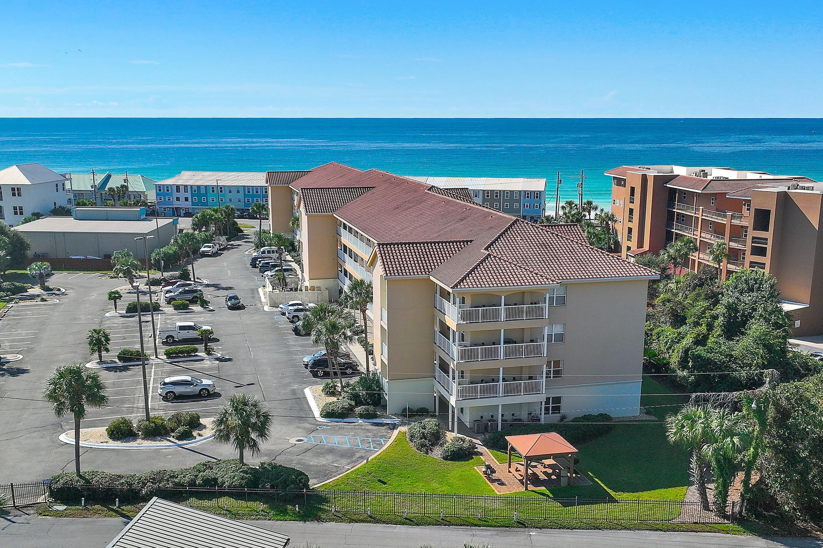 A multi-story hotel with a parking lot, a small lawn area, and a view of the ocean.