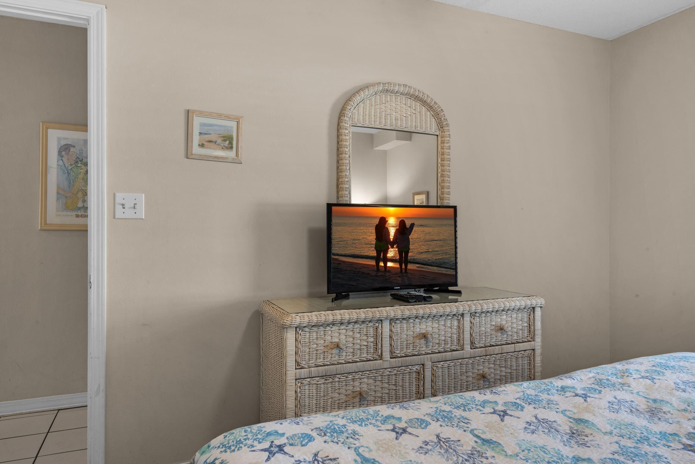 Un dormitorio con una televisión mostrando una escena de atardecer, un aparador de mimbre, una cama con colcha estampada y un espejo sobre el aparador.