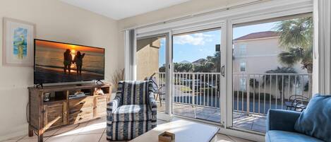 A living room with a television displaying a sunset scene, a wooden cabinet, a patterned armchair, a blue sofa, and a sliding glass door leading to an outdoor area.
