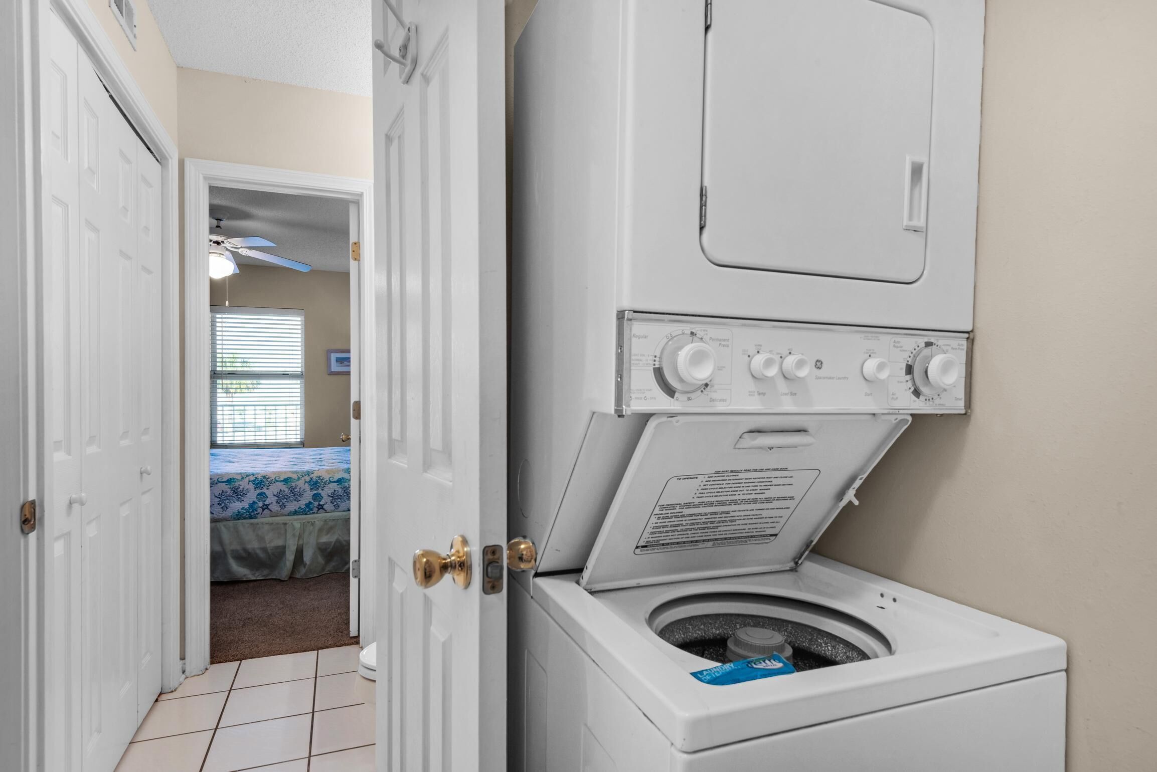 A laundry room with a front-loading washer and dryer, a door leading to a bedroom with a bed and a ceiling fan, and a tiled floor.