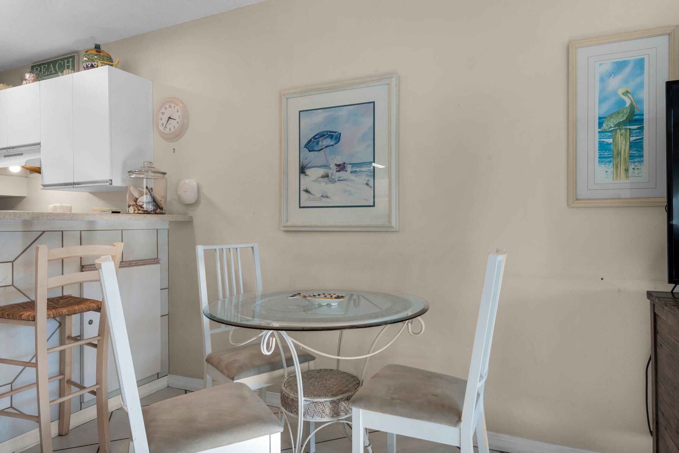 A dining area with a glass table and white chairs, a clock, and a framed beach painting on the wall.