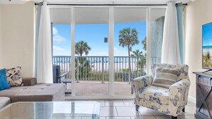 Ein Wohnzimmer mit einem Glastisch, einer Couch, einem gemusterten Sessel und Blick auf einen Strand durch eine Glasschiebetür.