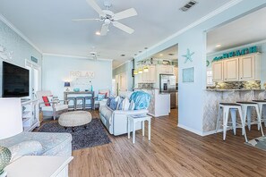 A spacious living room with a large TV, a ceiling fan, and a kitchen area with white cabinets and a bar stool. - Heart and Sol (Destin)