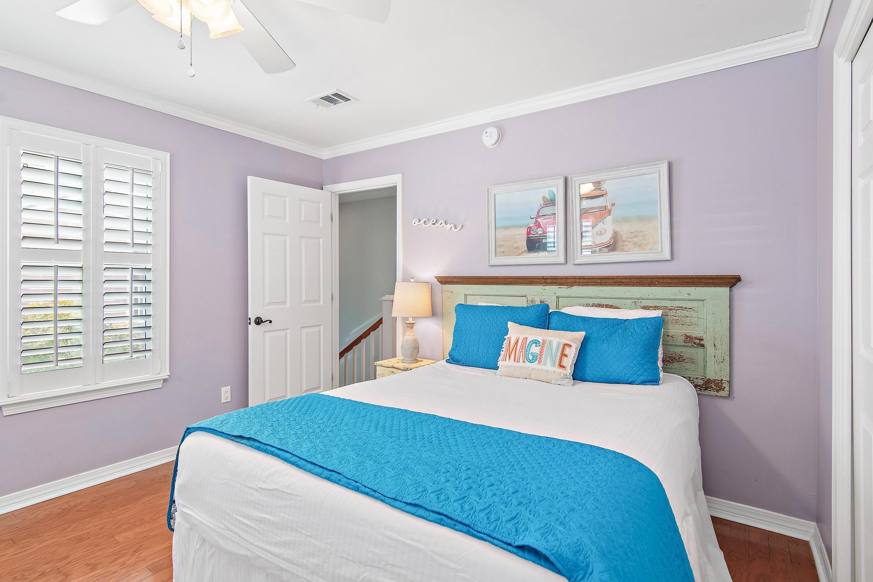 A bedroom with a bed, a door, a window with shutters, a ceiling fan, and a wall with two framed pictures.