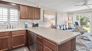A kitchen with wooden cabinets, a granite countertop, and a dishwasher. There is a dining area with a sofa and a chair, and a window with curtains.