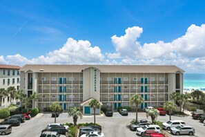 A multi-story hotel with a central entrance, parking lot, and a view of the beach.