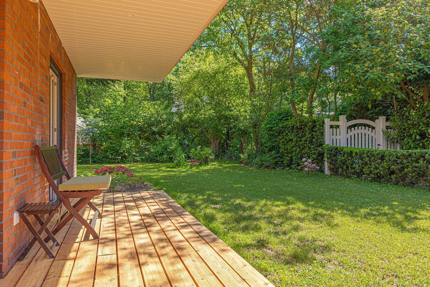 A wooden deck with a table and chairs, surrounded by a lush garden and a brick wall.