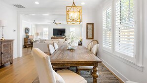 A dining area with a wooden table, white chairs, and a chandelier. There is a TV in the background and a window with blinds. - Once Upon a Tide (Miramar Beach)