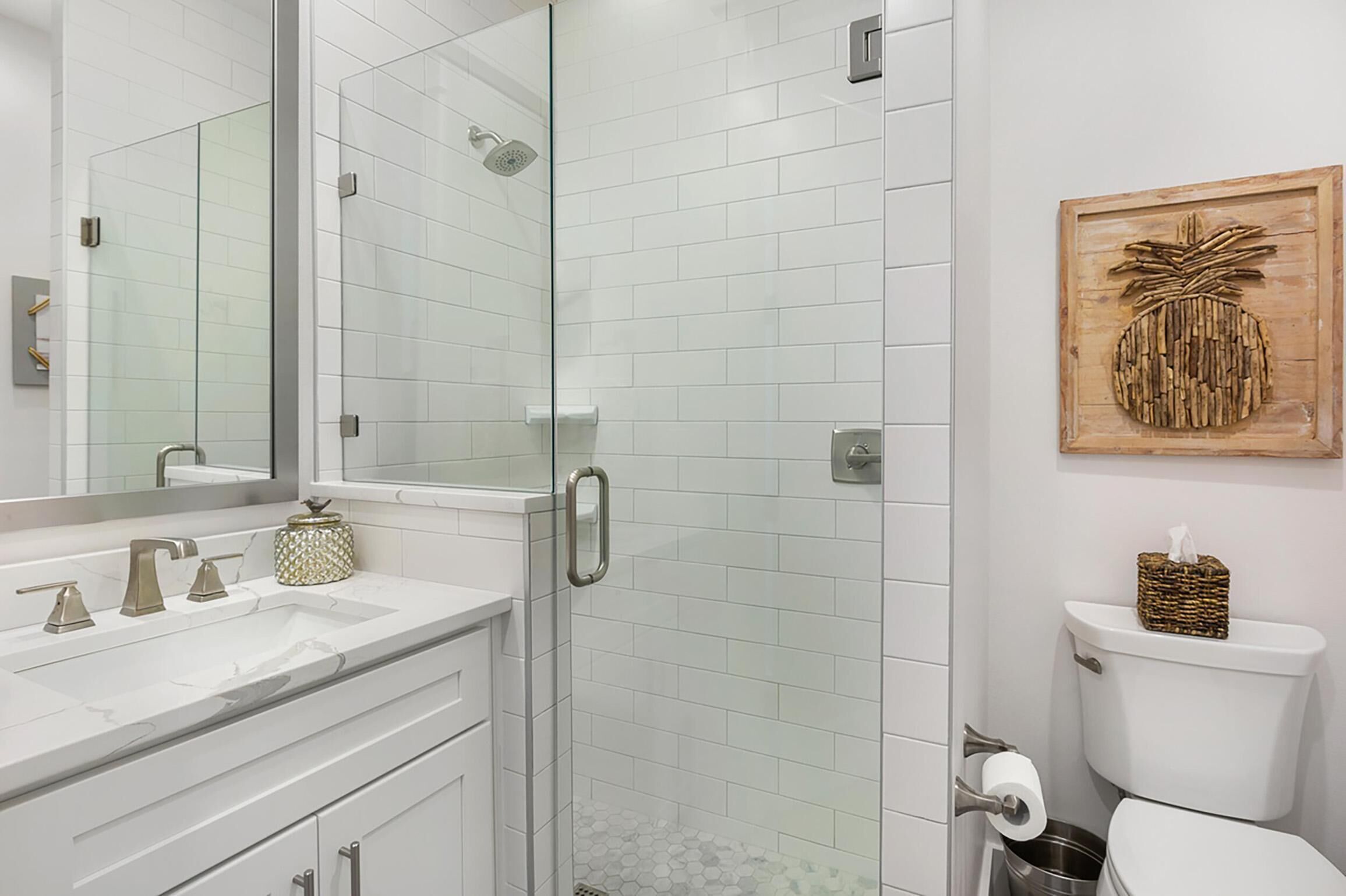 A modern bathroom with a glass-enclosed shower, white tiled walls, a large mirror, a sink with two faucets, and a toilet with a decorative basket on top.