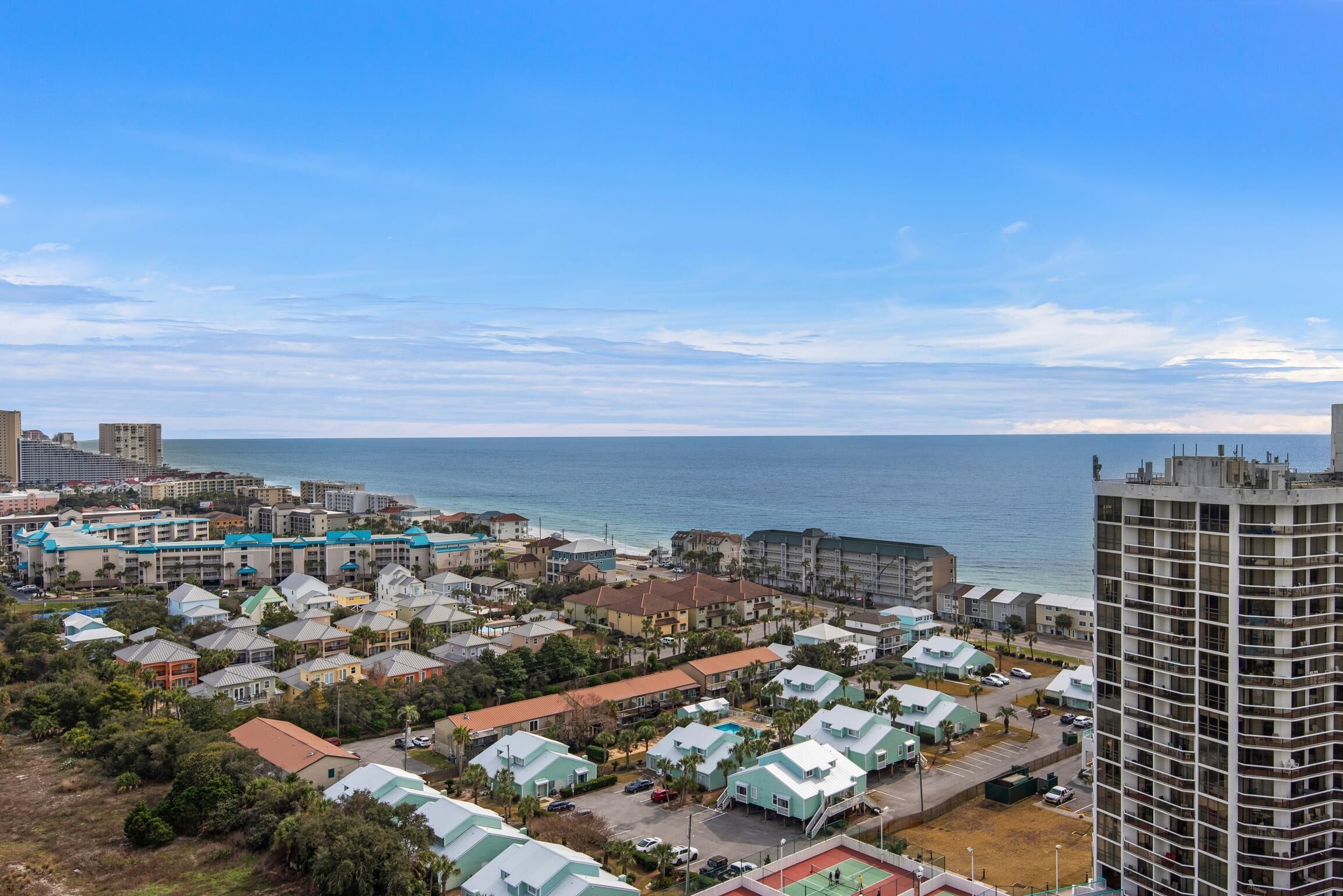 A coastal residential area with a mix of single and multi-story buildings, a clear view of the ocean, and a parking lot.