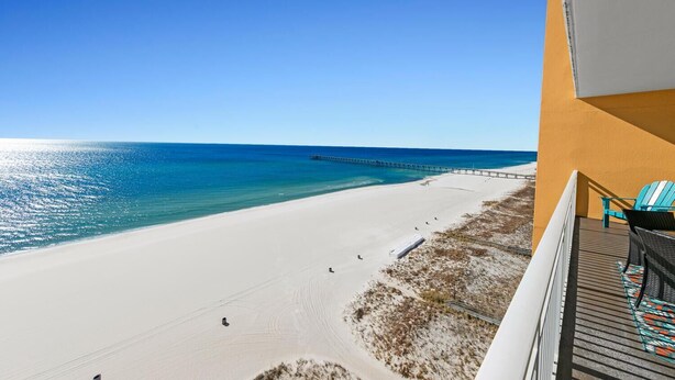 A beach view from a balcony with a wooden deck and a chair, overlooking a sandy beach and a pier extending into the ocean.