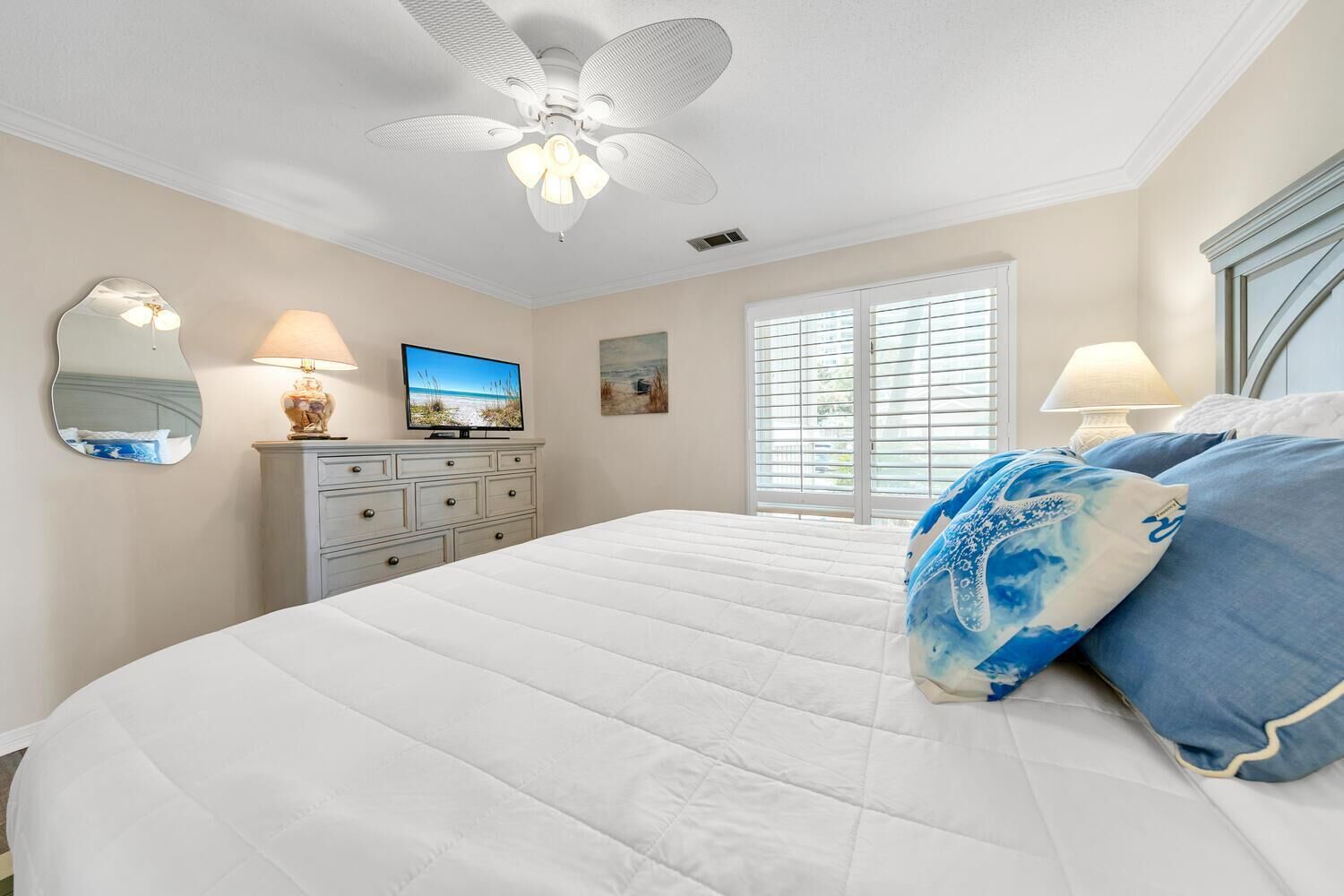 A bedroom with a large bed, a ceiling fan, a television, a dresser, and a window with blinds.