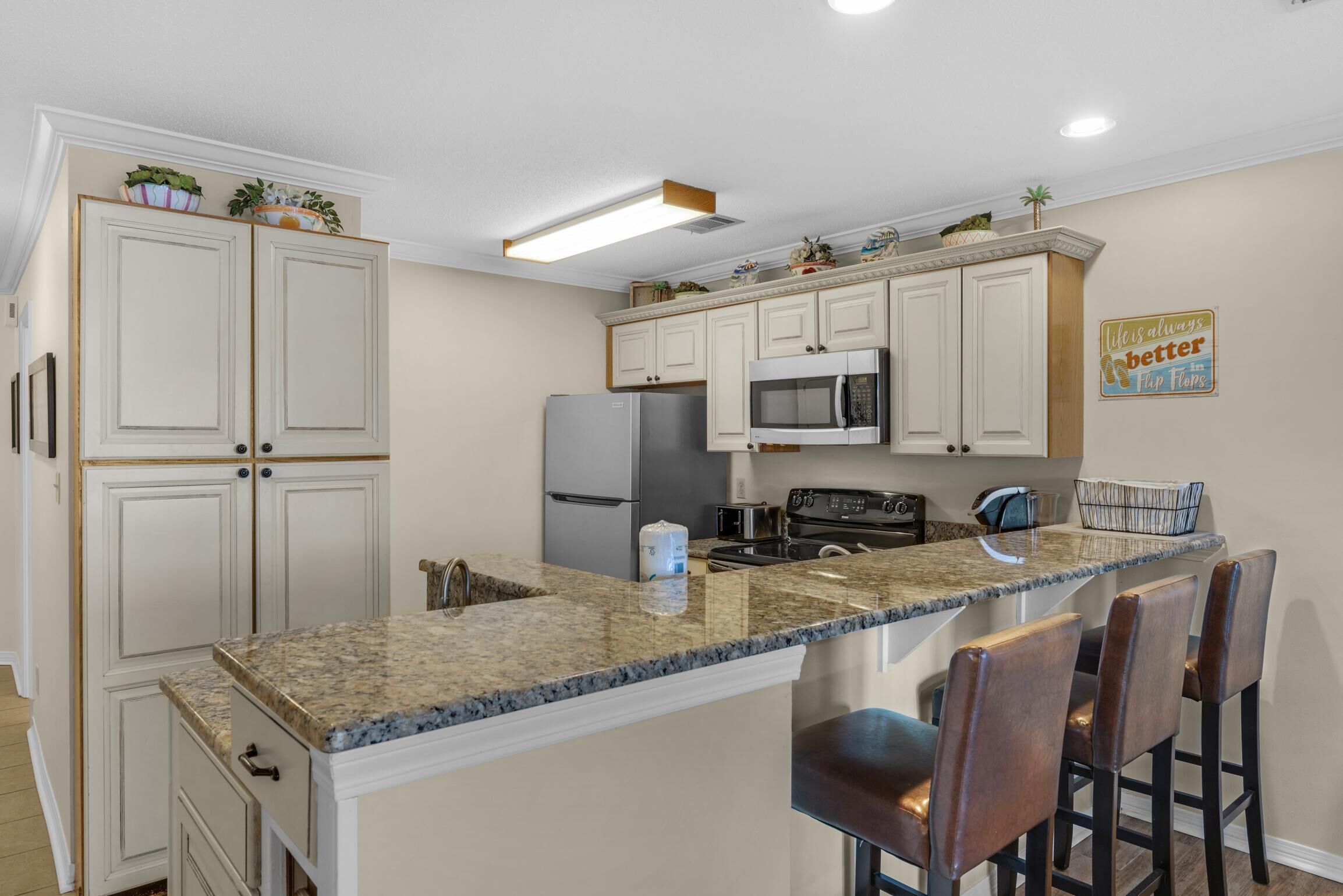 A kitchen with white cabinets, granite countertops, a refrigerator, microwave, and a coffee maker. There are bar stools and a sign on the wall.