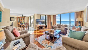 A spacious living room with a large TV, comfortable seating, a dining area, and a view of the ocean. - Edgewater Tower 1-411 (Panama City Beach)