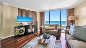 A living room with a large window, a sofa, a coffee table, and a television displaying a beach scene. - Edgewater Tower 1-411 (Panama City Beach)