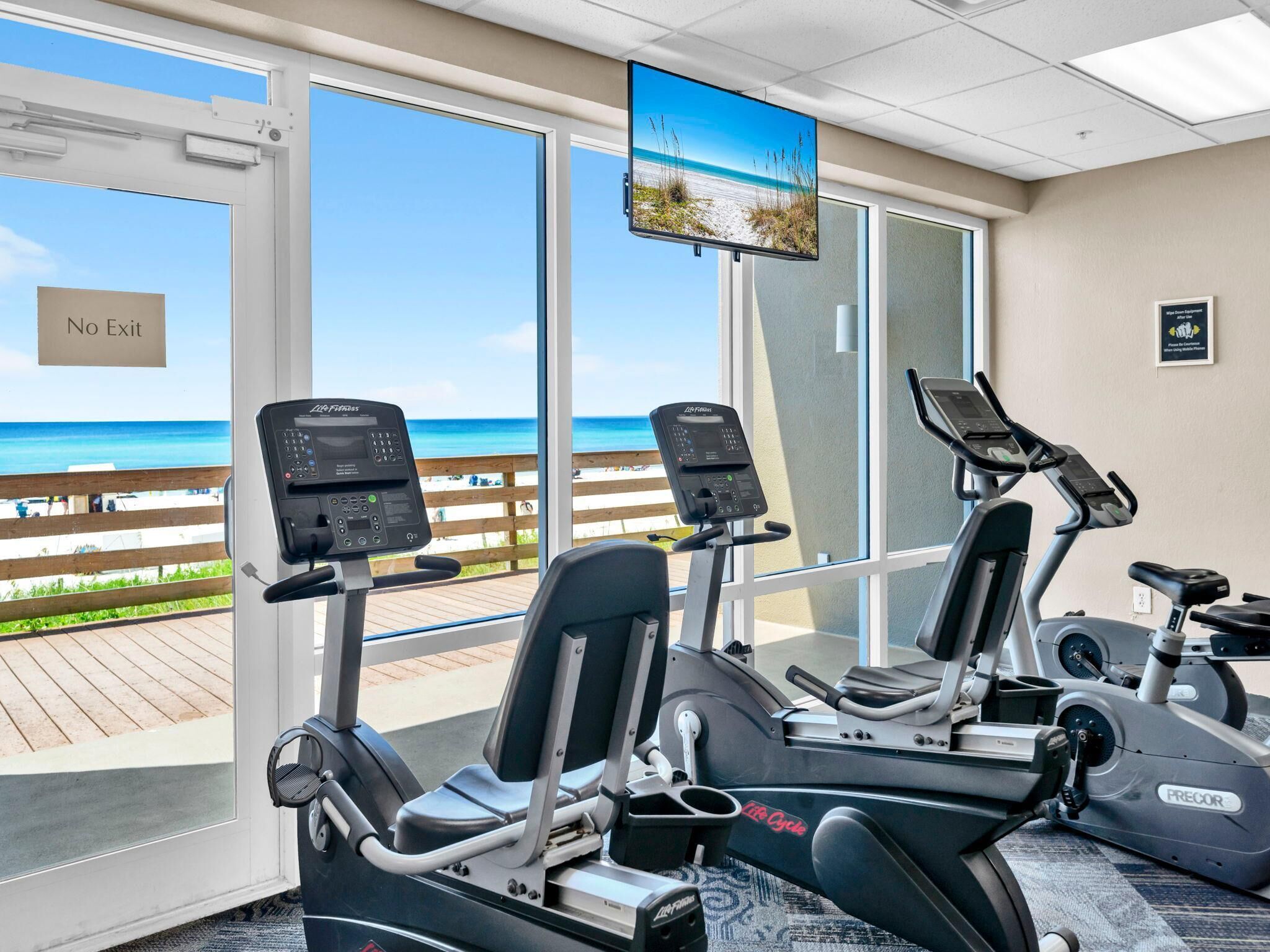 Un gimnasio con máquinas de cardio, una pantalla de televisión y vista a la playa.