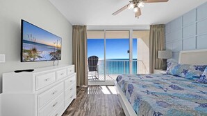 A bedroom with a large bed, a TV on the wall, a ceiling fan, and a balcony with a view of the ocean.