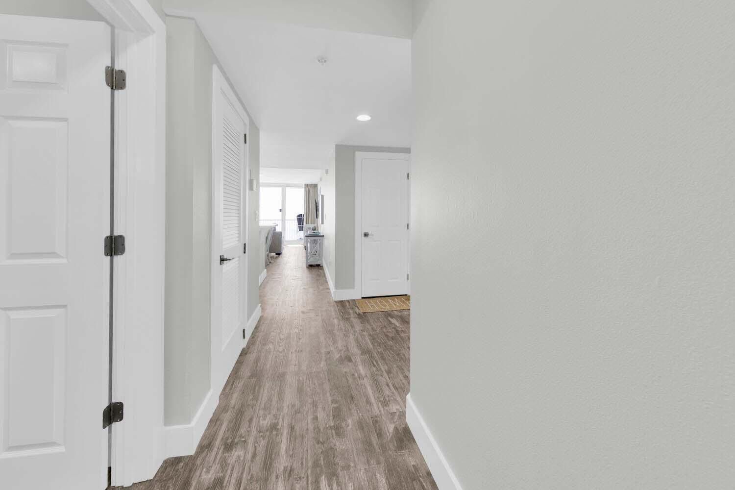 Un pasillo con paredes blancas y suelo de madera, que conduce a una habitación con ventana y puerta.