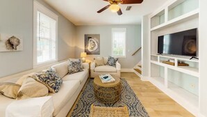 Un salon avec un canapé blanc, une table basse ronde en osier, un ventilateur de plafond et un téléviseur posé sur un meuble blanc.