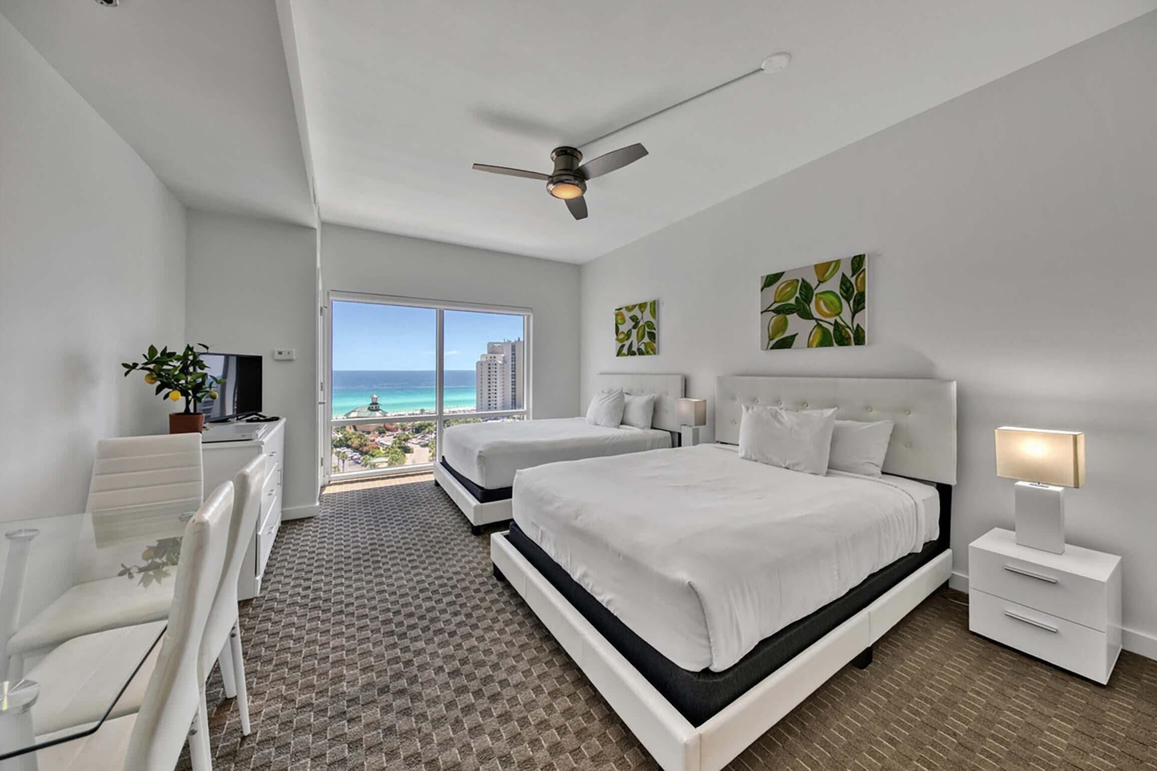A modern hotel room with two beds, a desk, and a view of the ocean.