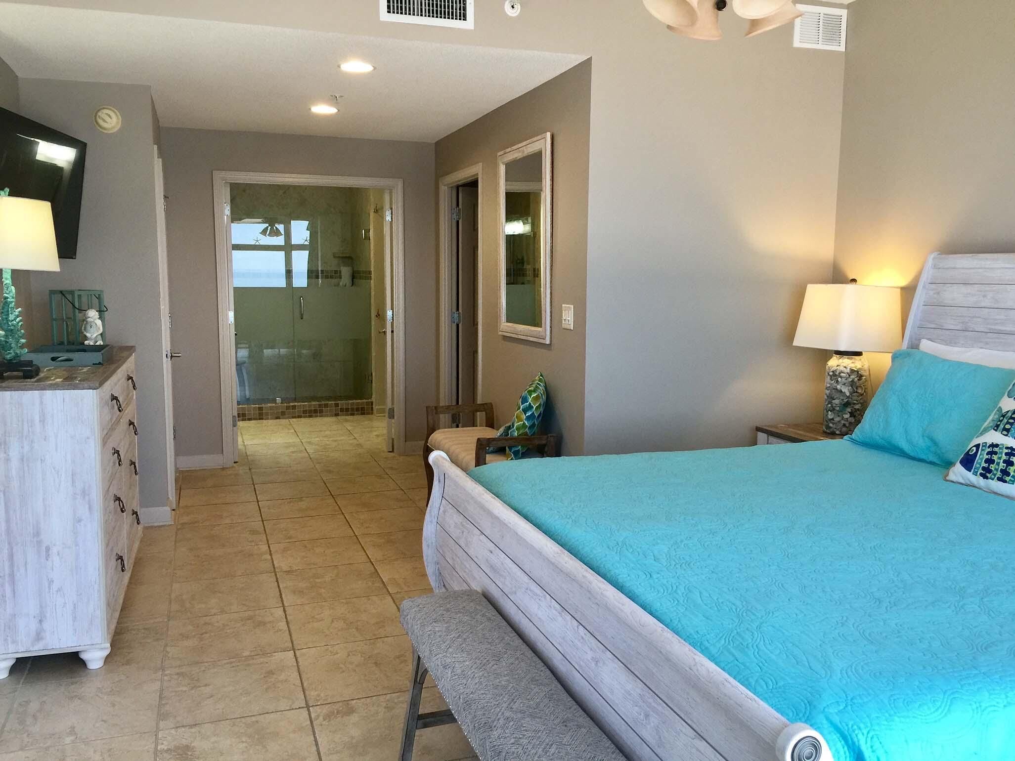 A bedroom with a bed, a bench, a dresser, a mirror, and a shower area.