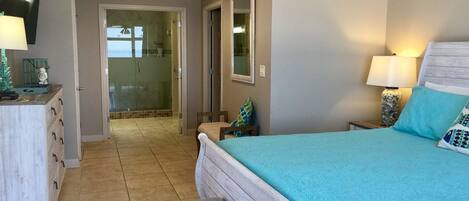 A bedroom with a bed, a bench, a dresser, a mirror, and a shower area.