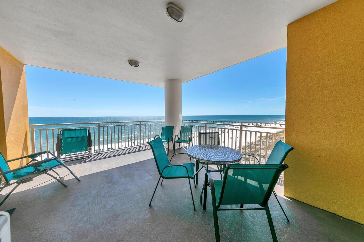 A balcony with a view of the beach, green chairs, and a round table.