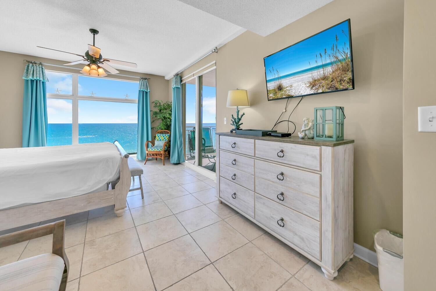A room with a bed, a dresser, a ceiling fan, a TV, and a view of the ocean.