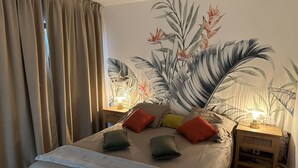 A bedroom with a bed, pillows, a nightstand, a lamp, and a wall mural. - La Voile du Rocher (Nouméa)