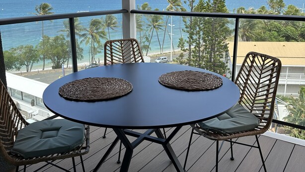 A round table with two woven placemats, surrounded by wicker chairs with cushions, on a wooden deck with a view of the ocean and palm trees. - La Voile du Rocher (Nouméa)
