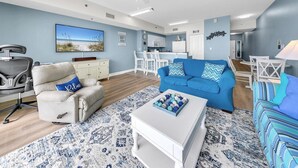 Uma sala de estar moderna com um sofá azul, uma poltrona cinza e uma mesa de centro branca. Ao fundo, dispõe-se uma área de cozinha com armários brancos e uma mesa de jantar.