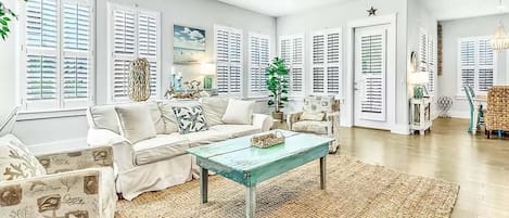 A spacious living room with a light-colored sofa, a coffee table, and a large window with shutters.