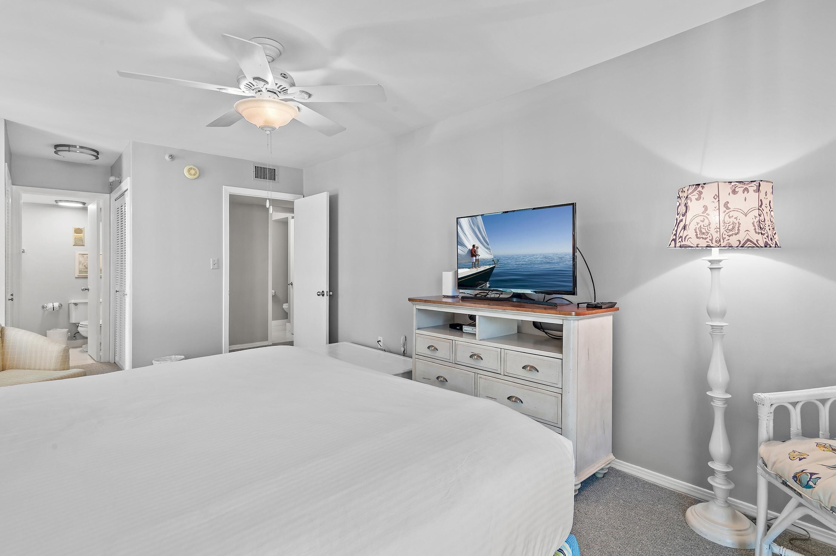 A bedroom with a large bed, a ceiling fan, a TV on a wooden stand, a lamp, and a chair.