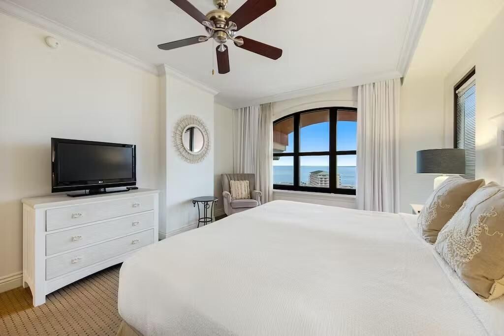Una camera d'albergo con un letto, una TV su un comò, una ventola a soffitto, vista sull'oceano e una finestra con tende.