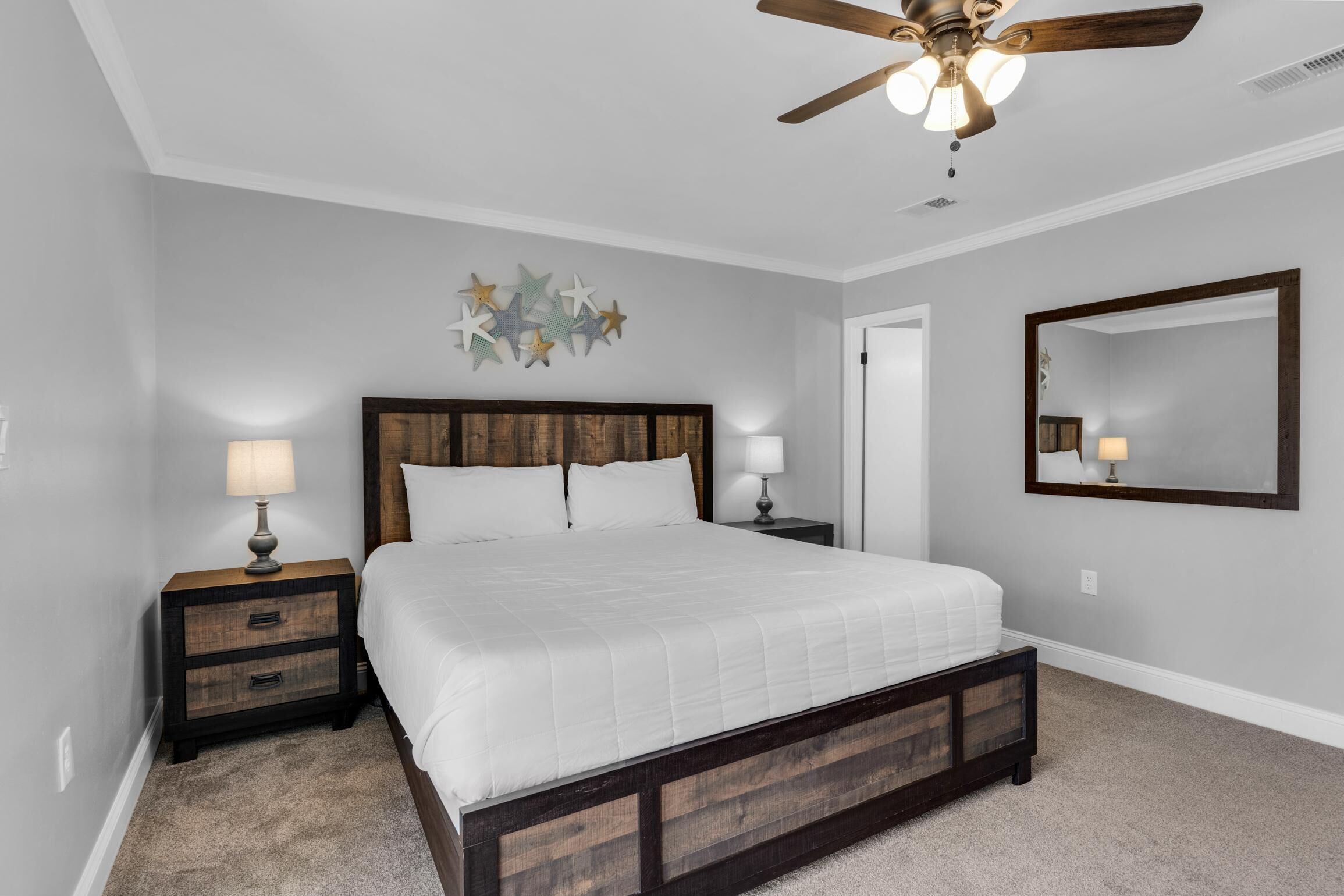Une chambre avec un grand lit, une tête de lit en bois, deux tables de chevet avec des lampes, un ventilateur de plafond, un miroir fixé au mur et une décoration murale en forme d’étoile.