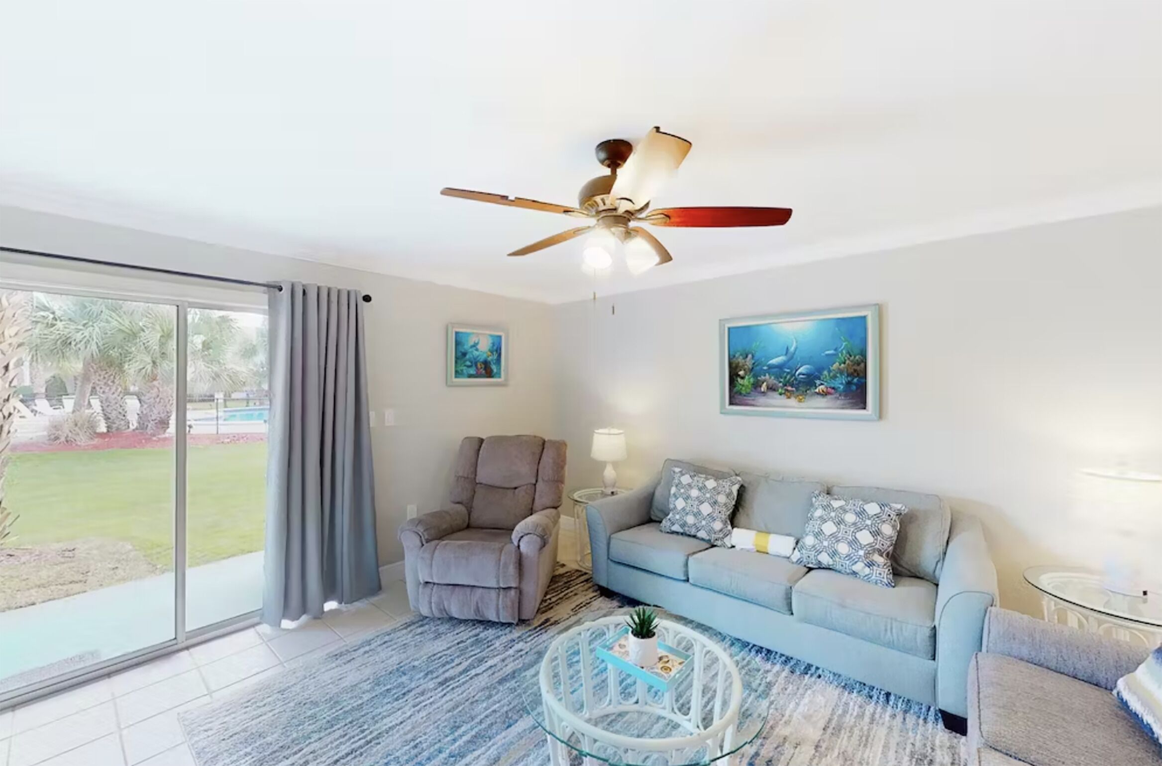 A living room with a ceiling fan, a light-colored sofa, a chair, a coffee table, and a large window with curtains.
