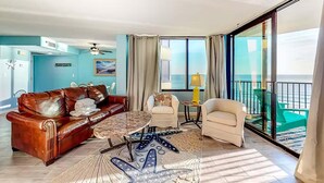 A spacious living room with a brown leather sofa, a round coffee table, and a beige armchair. A large window offers a view of the ocean.