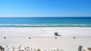 A wide sandy beach with a small structure in the center, likely a lifeguard hut, and a clear blue sky.