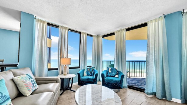 A spacious living room with a view of the beach, a round marble table, two blue armchairs, and a beige sofa.