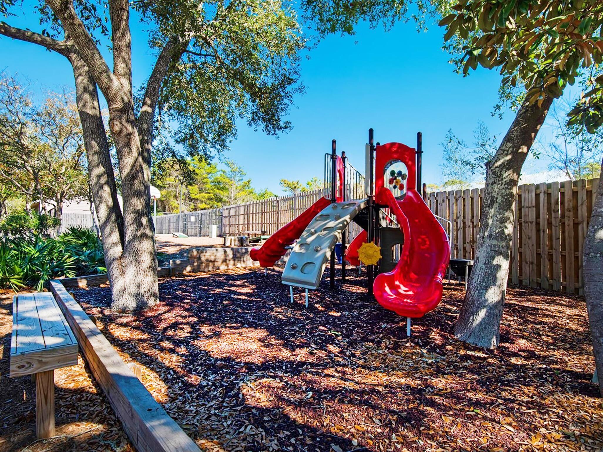A playground with a red slide and climbing structures, surrounded by trees and a wooden fence.