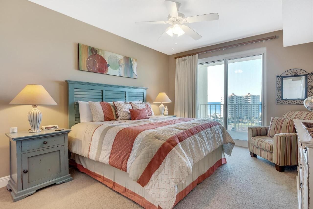 A bedroom with a bed, nightstands, a chair, a ceiling fan, and a large window with a view.