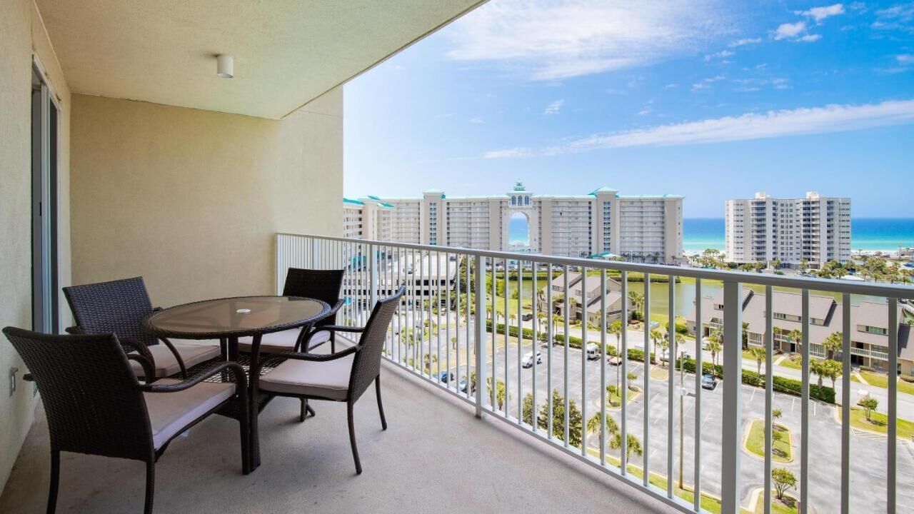A balcony with a table and chairs, overlooking a parking area and buildings with a clear view of the ocean.