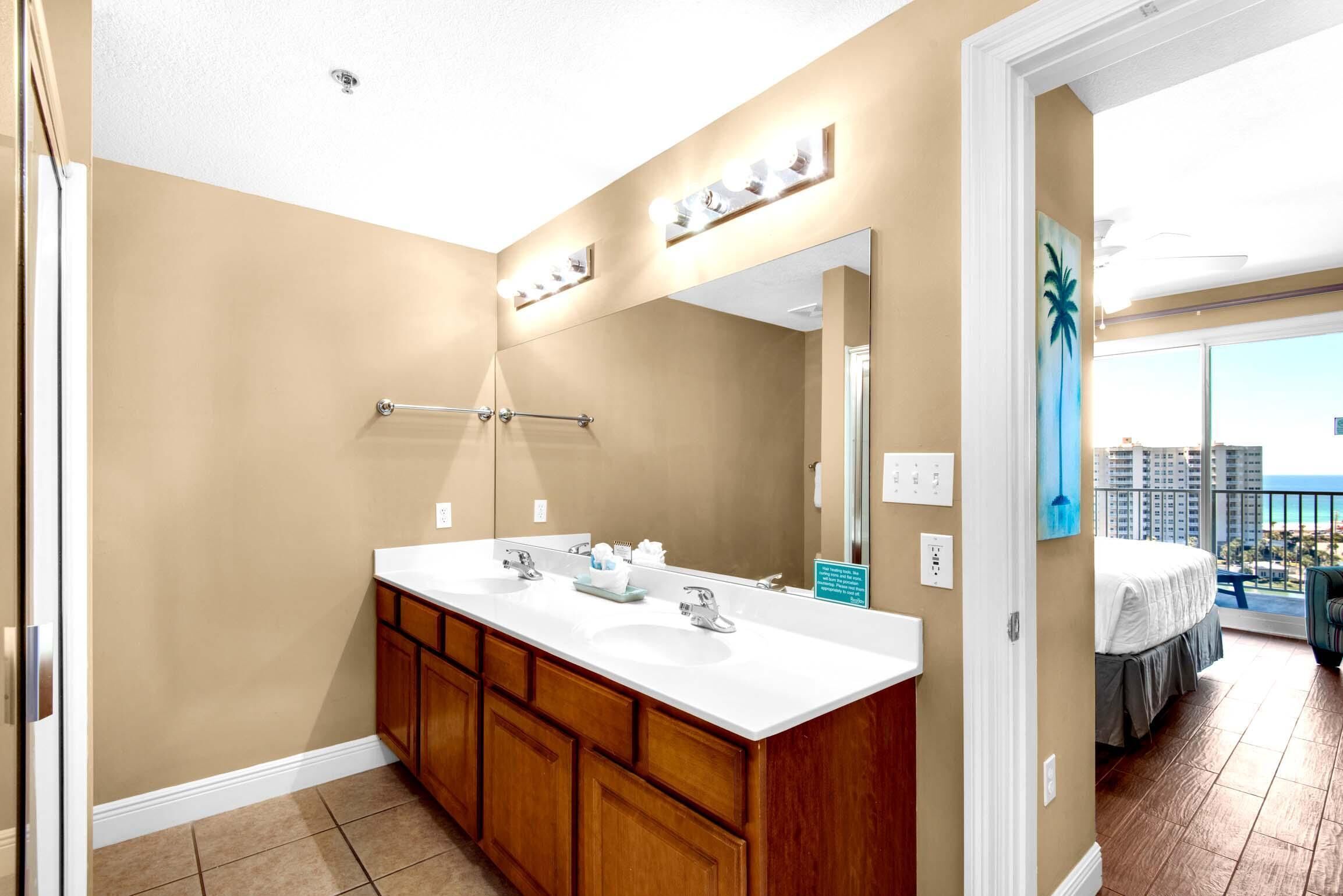 A bathroom with a double sink, wooden cabinets, a large mirror, and a view of a balcony with a bed.
