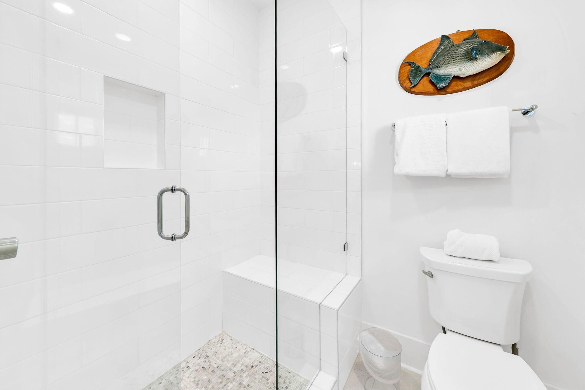 Ein modernes Badezimmer mit Duschkabine aus Glas, Toilette mit einer Holz-Fischdekoration und weiß gefliester Wand.