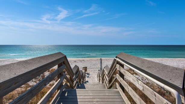 A wooden boardwalk leading to a beach with clear blue water and a bright sky.