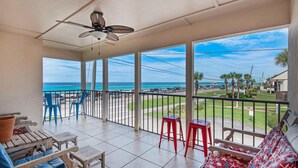 A balcony with a view of the ocean, blue chairs, red stools, and a ceiling fan.