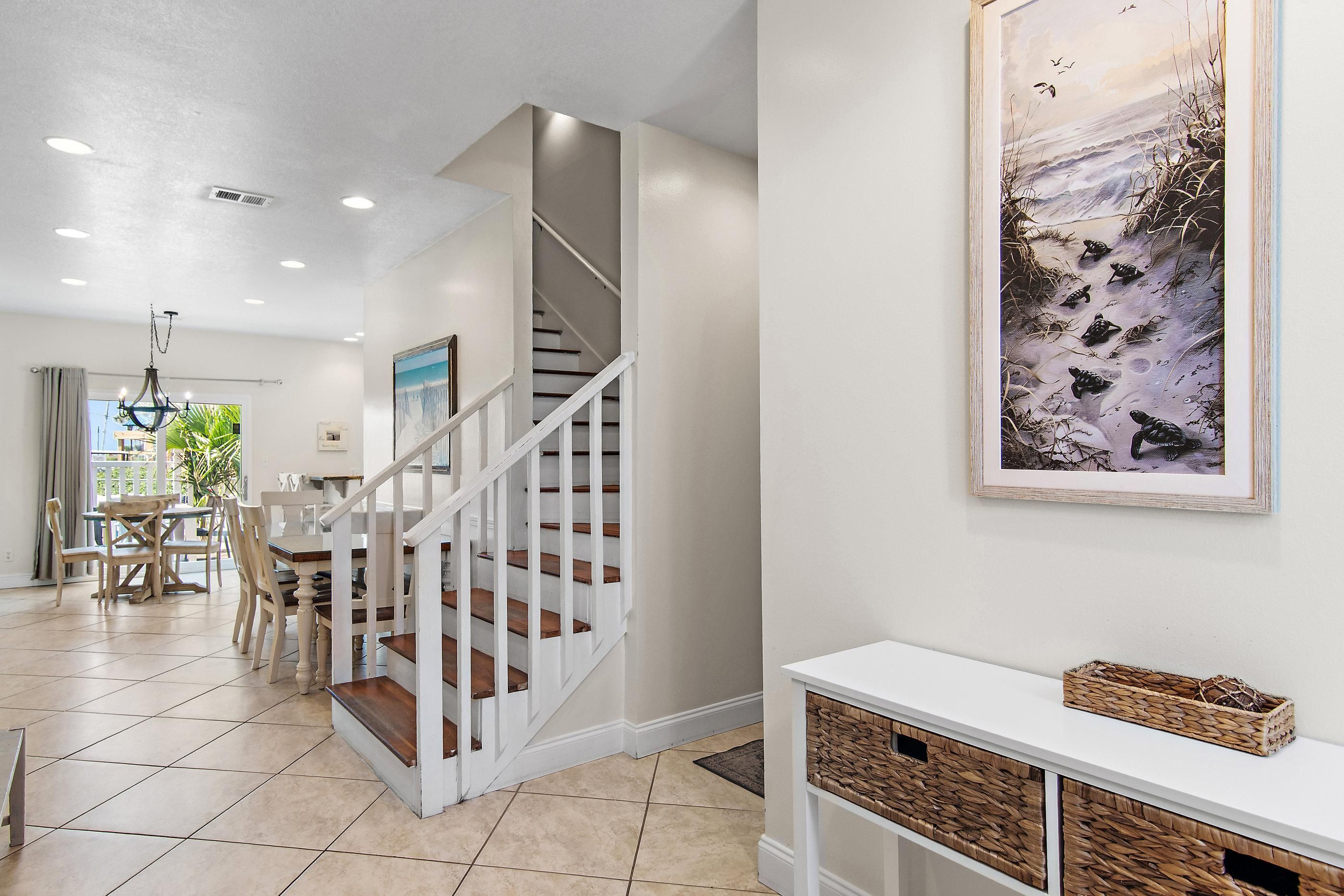 A staircase with white railings leading to an upper floor, a dining area with a table and chairs, and a framed artwork on the wall.
