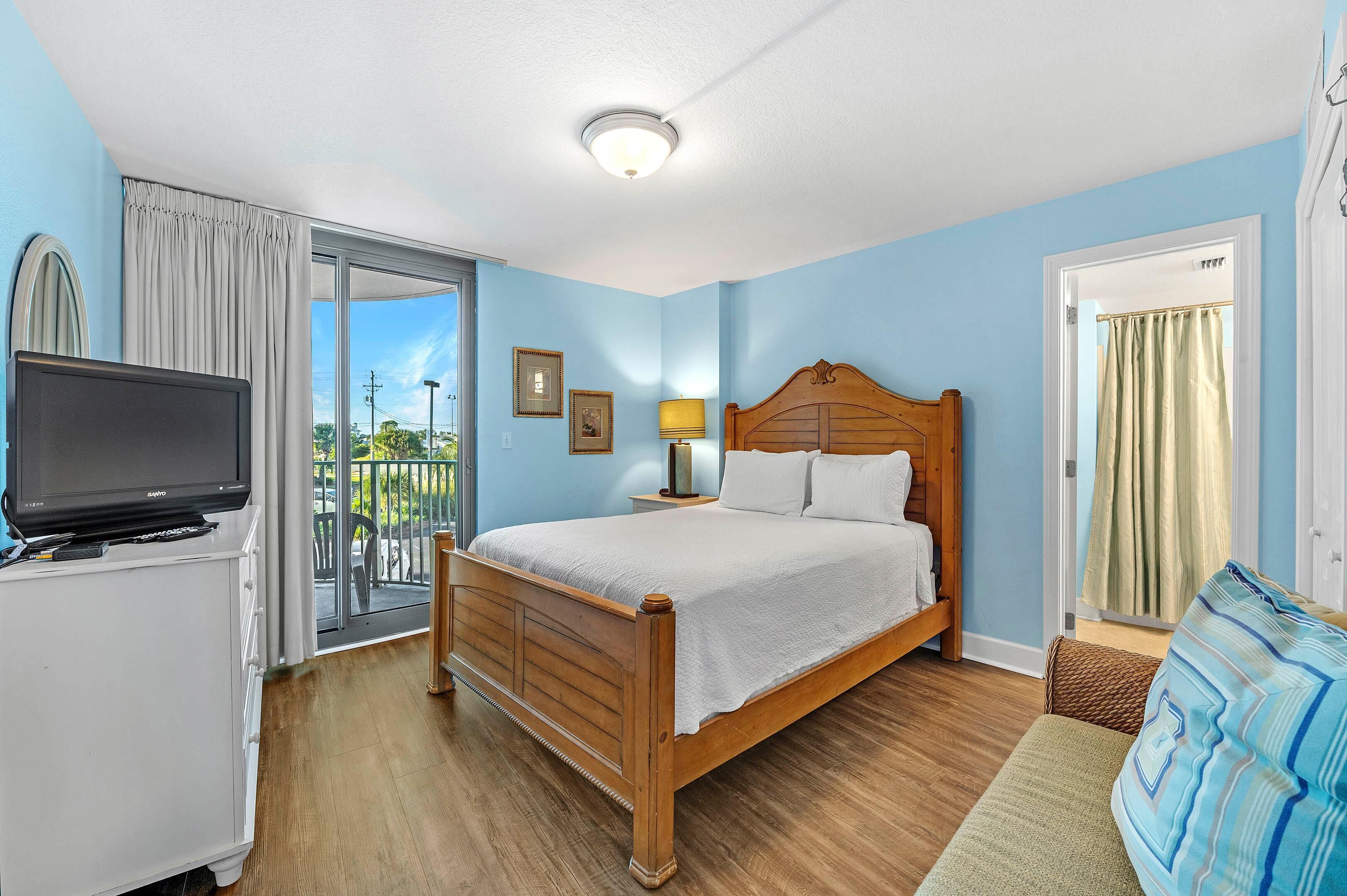 A bedroom with a wooden bed, a television on a white cabinet, a balcony with a view, and a door with curtains.