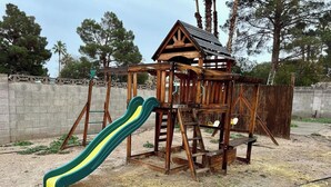Une structure de jeux en bois, dotée d’un toboggan et d’éléments d’escalade, entourée d’un mur en béton et d’arbres.